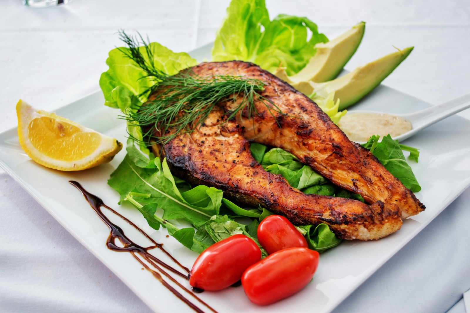 Grilovaný steak z lososa bohatý na omega-3 mastné kyseliny sedí na lůžku z křupavých listů salátu, doplněný plátky avokáda, plátkem citronu, malými rajčaty a hrstkou omáčky na bílém talíři. Roh talíře zdobí kapka balzamikové glazury.